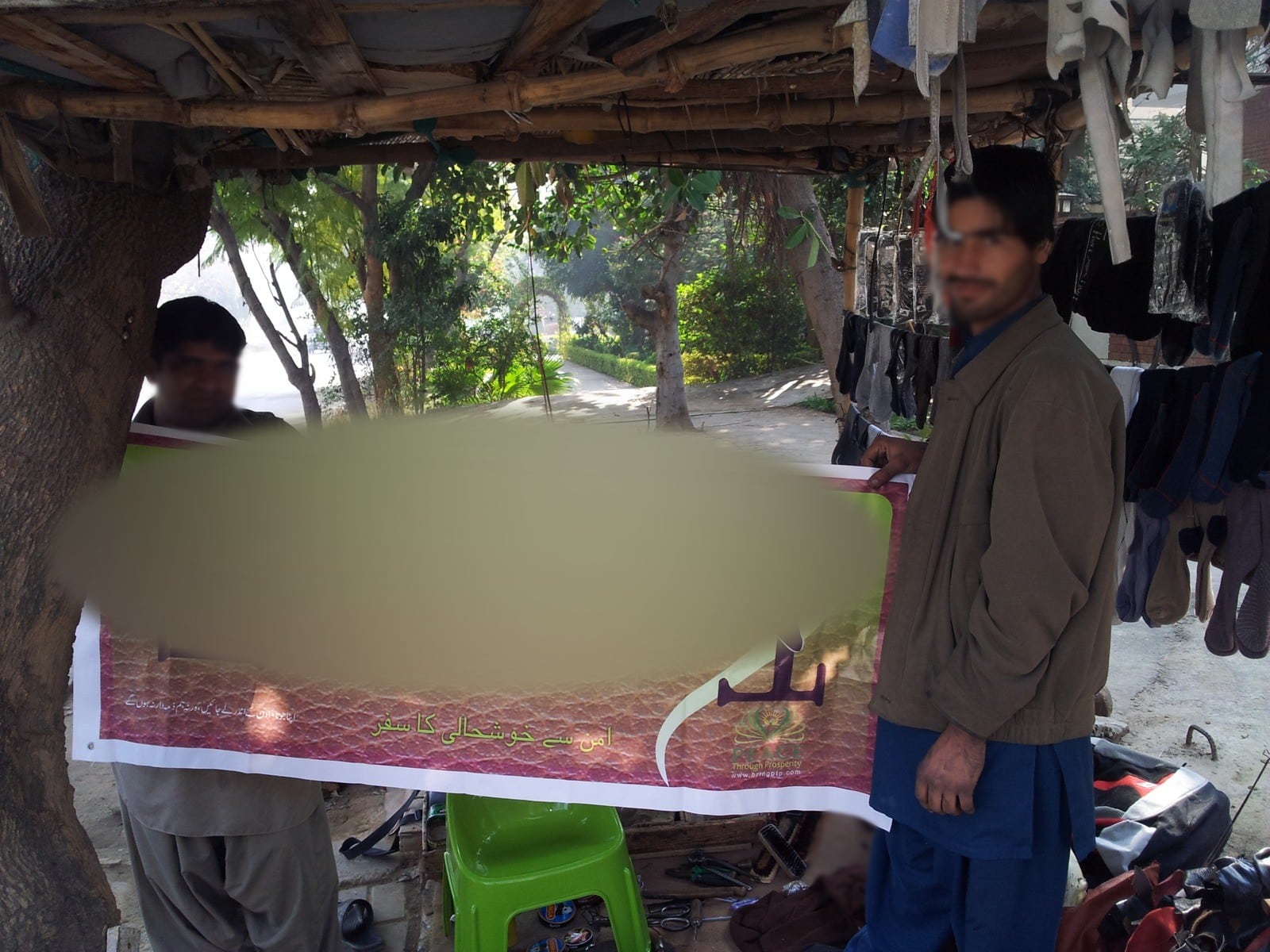 Two men holding up a new banner at a street cobbler's stall, Islamabad, 2013. Faces blurred.