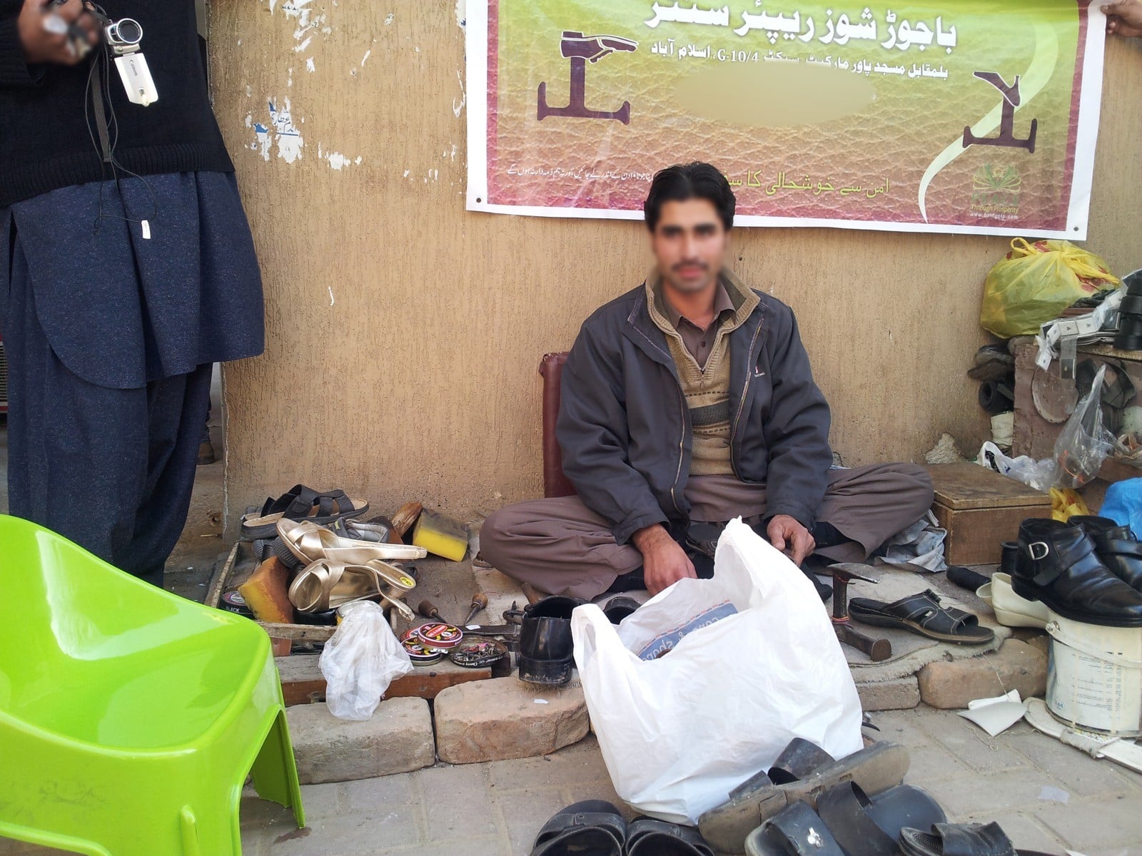 A street cobbler at his pitch in front of his new banner, Islamabad, 2013. Face blurred.