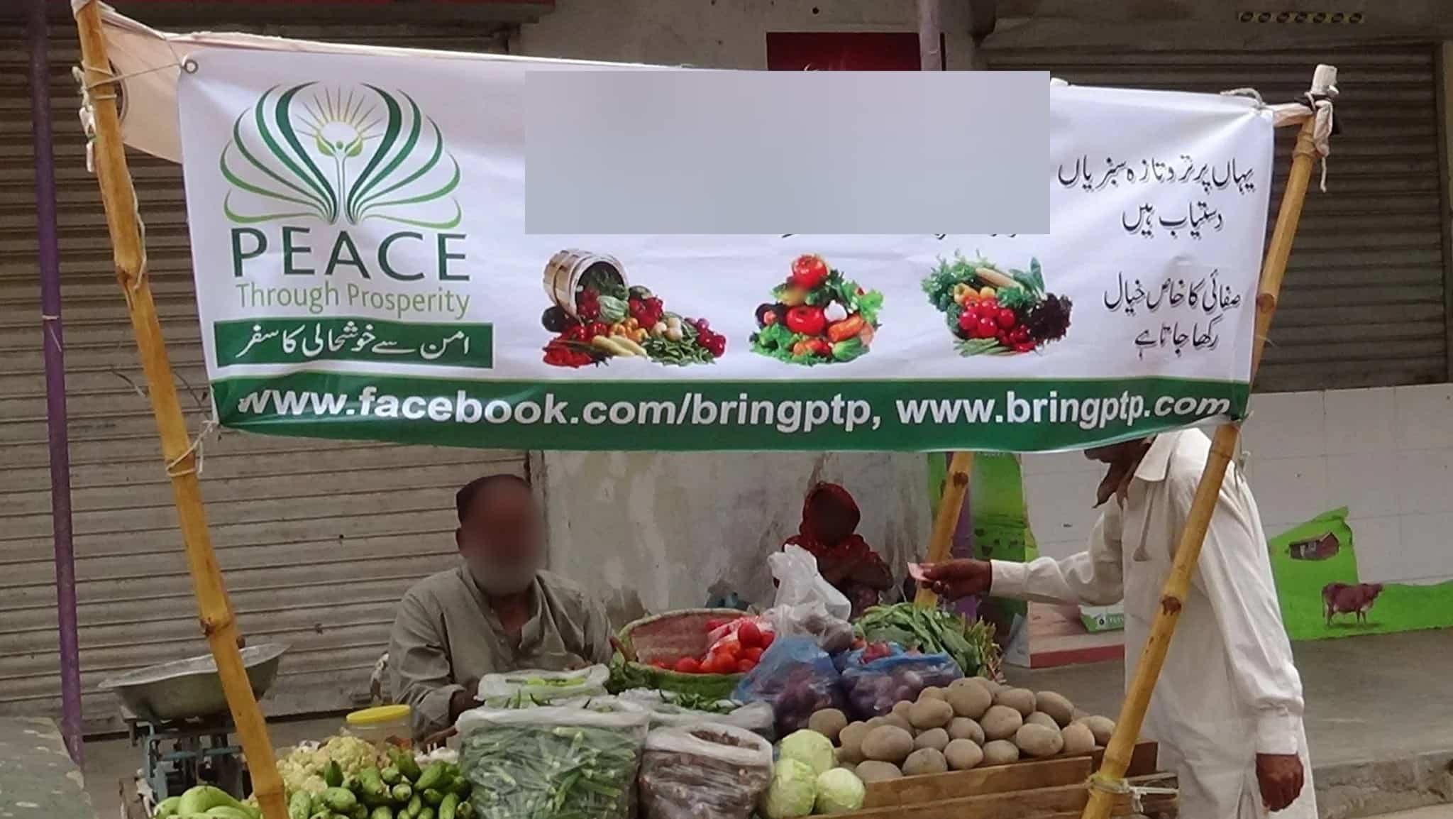 A vegetable stall in Karachi under a Peace Through Prosperity banner, with the stall name blurred for privacy