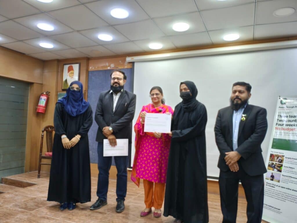 The two members of Scene Makers receiving a Certificate of Completion from Shahina Noor beside the programme standee at Sir Syed University, Karachi